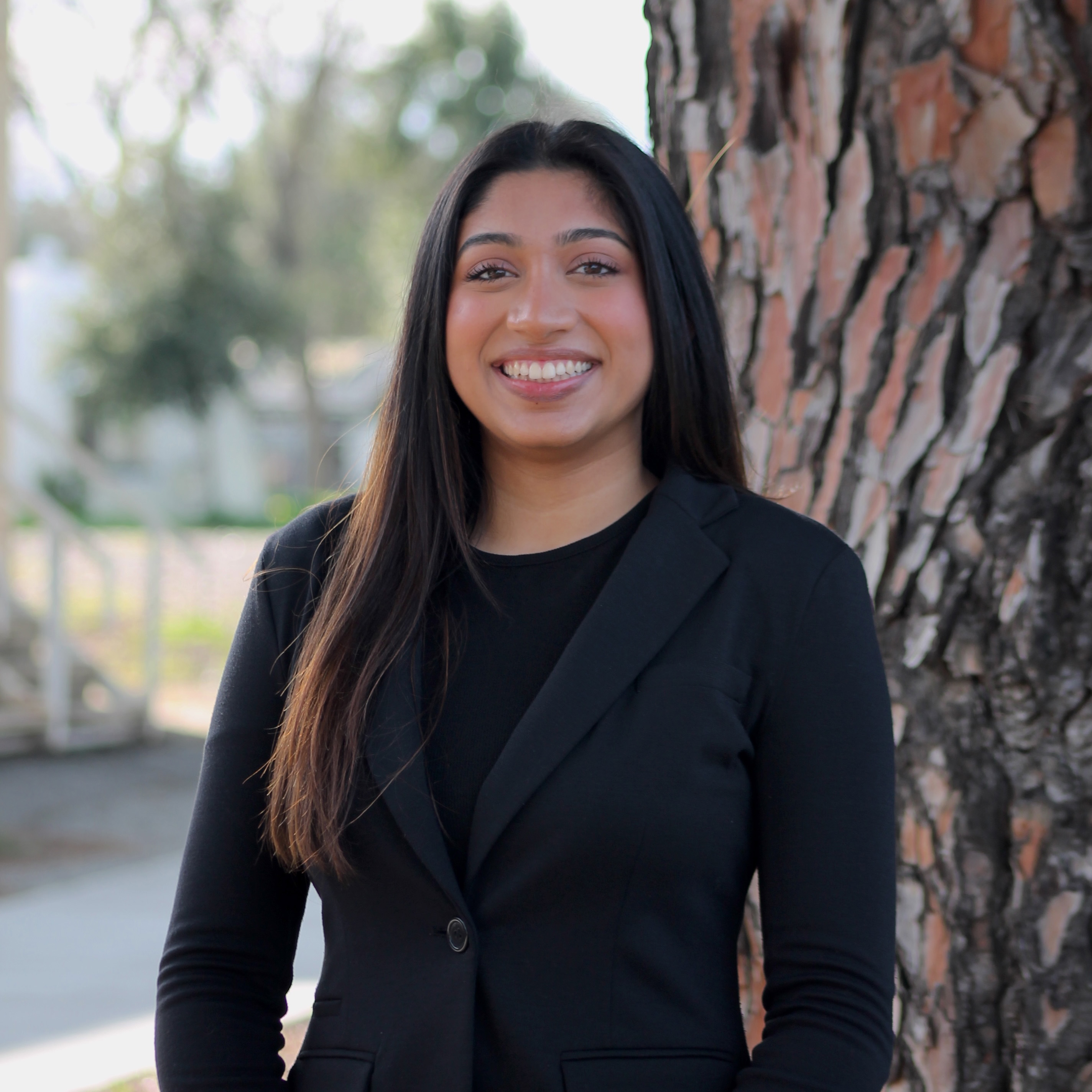 A young woman in a black suit smiles confidently against a tree backdrop.